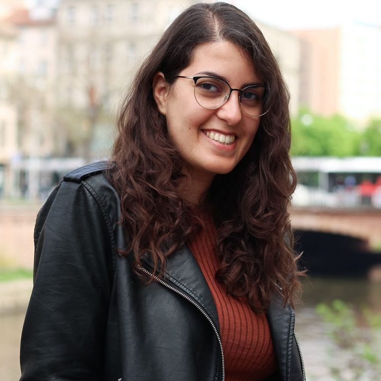 Femme souriante avec des lunettes, vêtue d'un manteau noir, devant un paysage urbain.