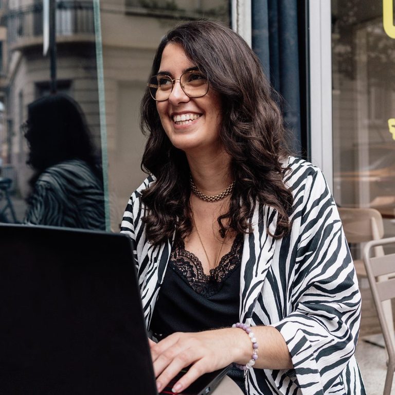Femme souriante avec des lunettes, vêtue d'un manteau noir, devant un paysage urbain.