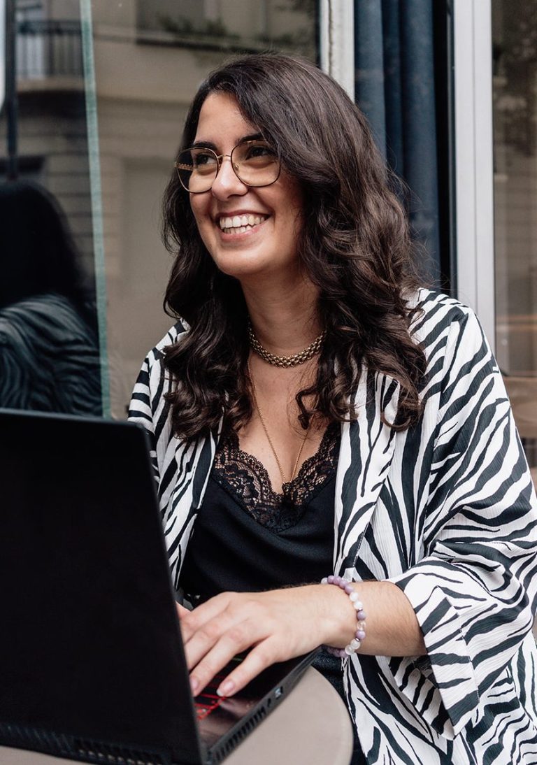 Pauline Fontaine-DOERR Femme souriante avec des lunettes, cheveux bouclés, devant un fond de fleurs.
